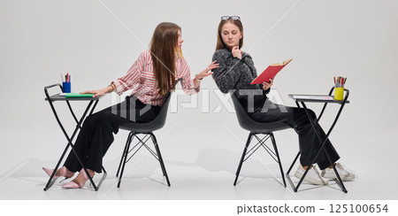 Teen girl leans to classmate from behind, attempting to distract her during lesson, communication against grey background. Teen girl leans to classmate from behind, attempting to distract her during lesson, communication against grey background. 125100654