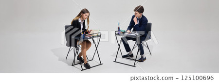 Two students, boy and girl in school uniforms, sitting at desk and studying together, making homework against grey studio background. 125100696