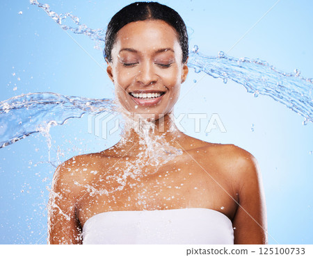 Woman, water splash and shower with body cleaning and smile with healthy skincare in blue studio background. Clean, face and water splash with an attractive young female cleaning in the bathroom 125100733