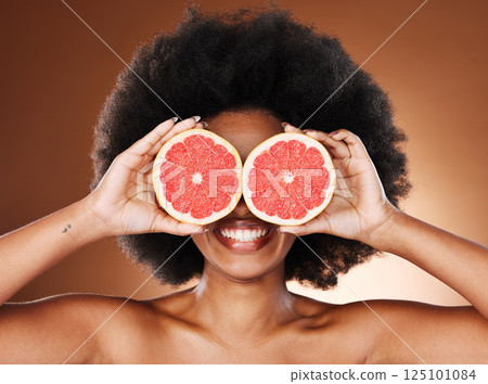 Beauty, skincare and black woman with grapefruit in hands for natural cosmetics and vitamin c facial treatment with studio background. Happy, smile and fruit covering eyes, wellness and healthy skin. 125101084