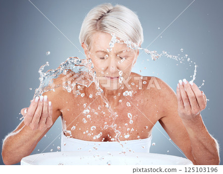 Senior woman, water splash and skincare with cleaning, wellness and face wash against a grey studio background. Elderly model, beauty and facials washing with liquid, hands and beauty treatment 125103196