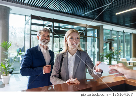 Businesswoman handing key card at reception desk while standing with male partner 125104167