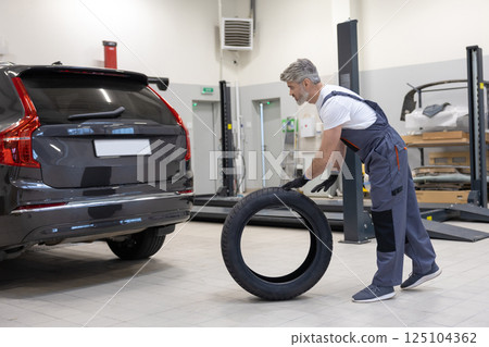Male mechanic in uniform holding new tire in auto workshop performing repair work Male mechanic in uniform holding new tire in auto workshop performing repair work 125104362
