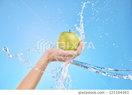 Hands, fruit and water splash for skincare nutrition, wellness or cosmetics against a blue studio background. Hand holding apple in healthy cleanse, hydration or hygiene for natural vitamin C skin 125104363