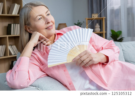 Smiling mid aged woman sitting on the sofa with a fan in hands 125105312