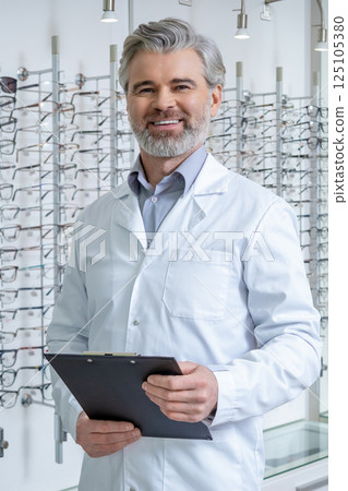 Mature doctor in lab coat standing near the stand with glasses and looking confident 125105380