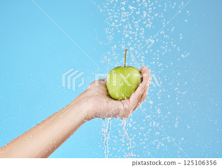 Green apple, hands and water splash for healthy lifestyle, vegan diet and nutrition on studio blue background. Palm holding fruits under clean running water for wellness, detox snack and vitamins 125106356