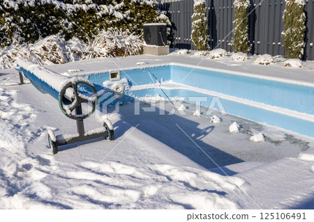 Outdoor swimming pool covered in snow and partially frozen in winter, with a pool cover roller also covered in snow. Backyard scene in cold weather. Outdoor swimming pool covered in snow and partially frozen in winter, with a pool cover roller also covered in snow. Backyard scene in cold weather. 125106491