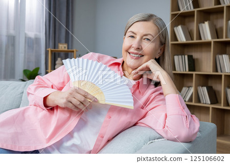 Smiling mid aged woman sitting on the sofa with a fan in hands 125106602