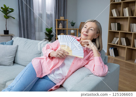 Smiling mid aged woman sitting on the sofa with a fan in hands 125106603