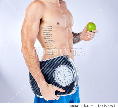 Body, scale and apple with a man in studio on a gray background for weightloss, diet or healthy eating. Fitness, tattoo and health with a male holding fruit while posing for a nutrition lifestyle 125107291