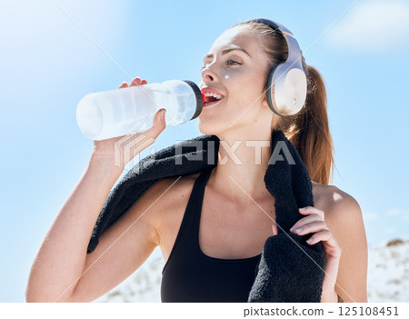 Woman, drink and water bottle during fitness, run outdoor with towel and music with headphones or streaming podcast for exercise motivation. Hydration, wellness and drinking water after workout. Woman, drink and water bottle during fitness, run outdoor with towel and music with headphones or streaming podcast for exercise motivation. Hydration, wellness and drinking water after workout. 125108451