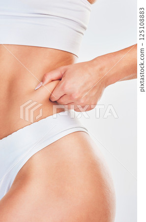 Hand, pinch and woman in underwear her belly fat for weight loss on a white studio background. Overweight, cellulite and fat with a woman with a healthy lifestyle and diet for stomach loss Hand, pinch and woman in underwear her belly fat for weight loss on a white studio background. Overweight, cellulite and fat with a woman with a healthy lifestyle and diet for stomach loss 125108883