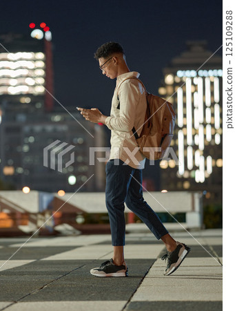 Night, man and phone while walking in a city with social media, networking and searching the internet. Male text contact, online and networking with internet contact on app with mobile smartphone Night, man and phone while walking in a city with social media, networking and searching the internet. Male text contact, online and networking with internet contact on app with mobile smartphone 125109288