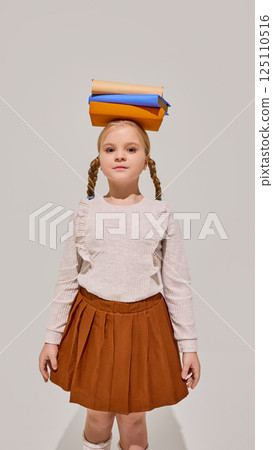 Young girl with braids stands confidently balancing stack of books on head, symbolizing knowledge, learning discipline, childhood curiosity. 125110516