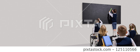 Kids standing at blackboard, solving problem using ruler while classmates watch attentively from desks against grey background 125110566