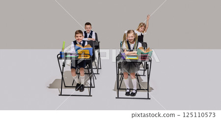 Four cheerful elementary school children sitting at desks in classroom, reading books, answering question on grey background. 125110573