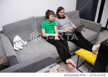 Cozy afternoon in a modern living space with mother and son engaged in technology and learning together while relaxing on a comfortable couch Cozy afternoon in a modern living space with mother and son engaged in technology and learning together while relaxing on a comfortable couch 125112328