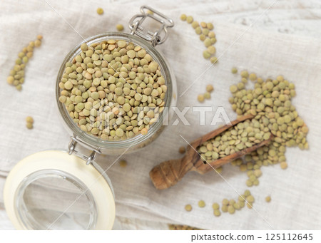 Glass jar full of raw green lentils with a wooden scoop on white table top view. Dried legumes 125112645