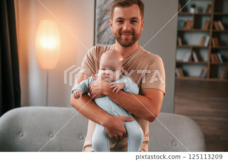 Cheerful mood. Holding in a hands. Father with his newborn baby is indoors. Conception of single dad Cheerful mood. Holding in a hands. Father with his newborn baby is indoors. Conception of single dad 125113209