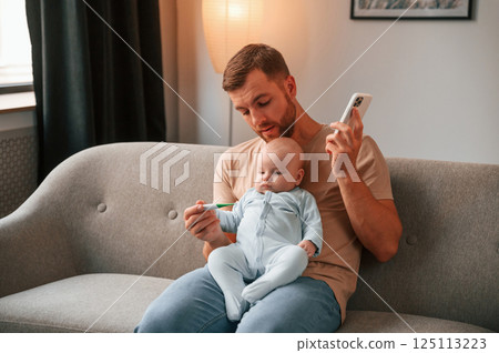 Sitting on the sofa. Father with his newborn baby is indoors. Conception of single dad Sitting on the sofa. Father with his newborn baby is indoors. Conception of single dad 125113223