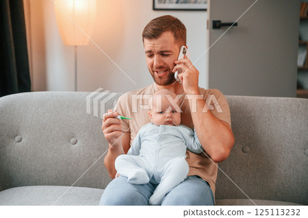 Calling doctor. With thermometer. Father with his newborn baby is indoors. Conception of single dad 125113232