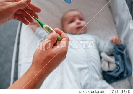 Holding thermometer, kid in the cradle. Father with his newborn baby is indoors. Conception of single dad Holding thermometer, kid in the cradle. Father with his newborn baby is indoors. Conception of single dad 125113258