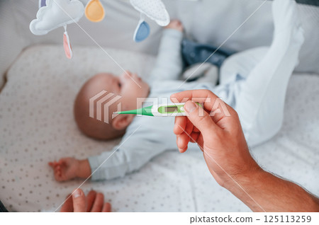 Holding thermometer, kid in the cradle. Father with his newborn baby is indoors. Conception of single dad Holding thermometer, kid in the cradle. Father with his newborn baby is indoors. Conception of single dad 125113259