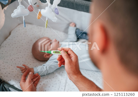 Holding thermometer, kid in the cradle. Father with his newborn baby is indoors. Conception of single dad Holding thermometer, kid in the cradle. Father with his newborn baby is indoors. Conception of single dad 125113260