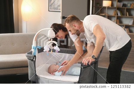Looking at the kid in cradle. Lovely couple taking care of newborn baby indoors at domestic room 125113382