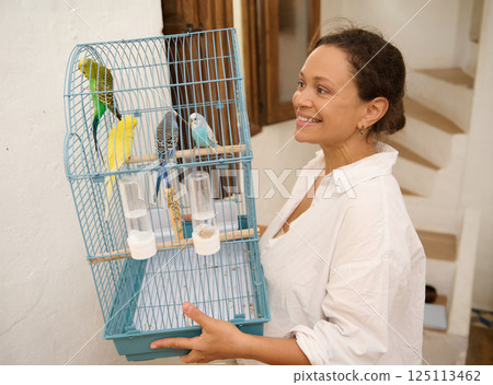 Smiling Woman Carrying a Bird Cage with Colorful Pet Parrots Indoors Smiling Woman Carrying a Bird Cage with Colorful Pet Parrots Indoors 125113462