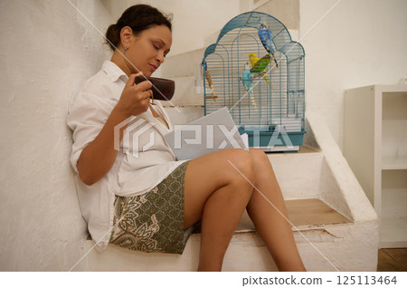 Serene Woman Reads While Relaxing Beside a Birdcage on a Stair Serene Woman Reads While Relaxing Beside a Birdcage on a Stair 125113464