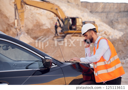Using laptop that is on the car and talking by phone. Man in uniform is working in the quarry at daytime Using laptop that is on the car and talking by phone. Man in uniform is working in the quarry at daytime 125113611