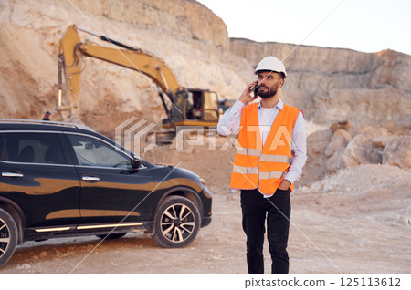 Standing near modern black automobile and talking by phone. Man in uniform is working in the quarry at daytime Standing near modern black automobile and talking by phone. Man in uniform is working in the quarry at daytime 125113612