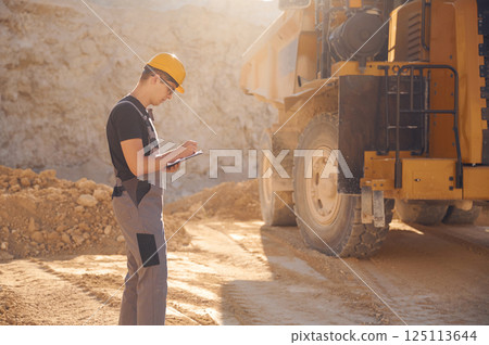 With documents near the haul truck. Man in uniform is working in the quarry at daytime 125113644