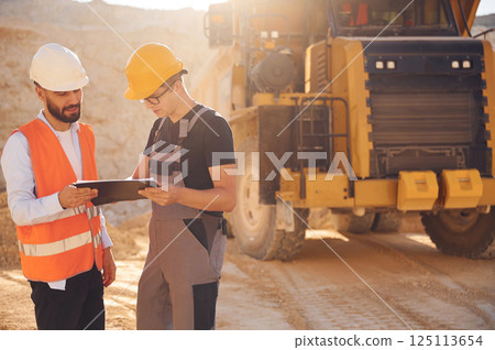 Standing near haul truck and talking about document. Two men in uniform are working together in the quarry Standing near haul truck and talking about document. Two men in uniform are working together in the quarry 125113654
