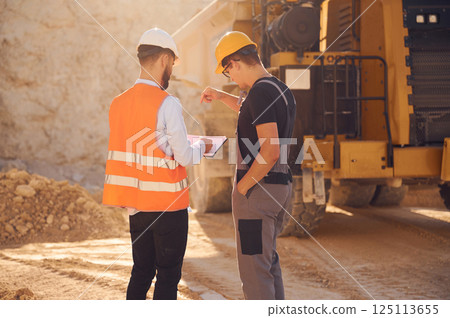 Standing near haul truck and talking about document. Two men in uniform are working together in the quarry Standing near haul truck and talking about document. Two men in uniform are working together in the quarry 125113655