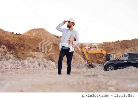 Touching hard hat and holding laptop. Man in uniform is working in the quarry at daytime 125113883