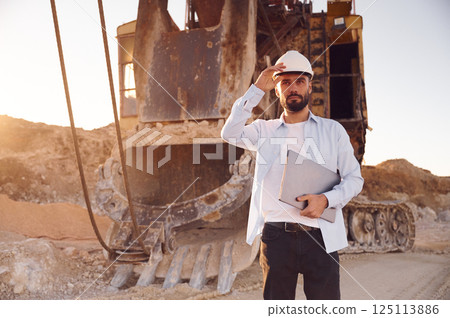 Standing against big excavator and holding laptop. Man in uniform is working in the quarry at daytime Standing against big excavator and holding laptop. Man in uniform is working in the quarry at daytime 125113886