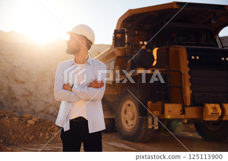 Standing against big haul truck. Man in uniform is working in the quarry at daytime Standing against big haul truck. Man in uniform is working in the quarry at daytime 125113900
