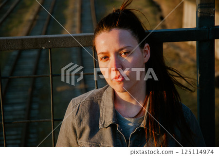 Thoughtful young woman on bridge at sunset with railway background 125114974