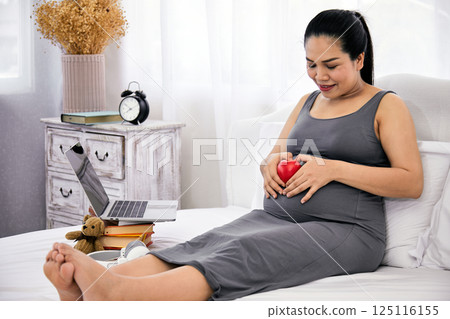 Aisan pregnant woman relaxing on bed smiling at red heart-shaped object on belly, surrounded by laptop, books, teddy bear and headphones in cozy bedroom 125116155