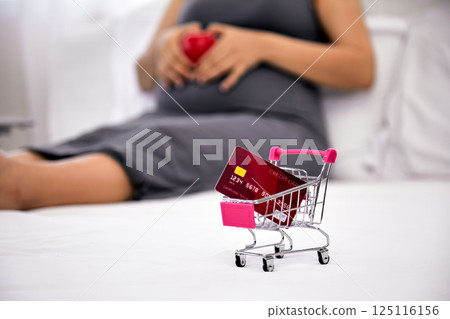 Mini shopping cart with credit card on white bed in front of pregnant woman holding red heart, online maternity shopping and financial planning during pregnancy at home Mini shopping cart with credit card on white bed in front of pregnant woman holding red heart, online maternity shopping and financial planning during pregnancy at home 125116156