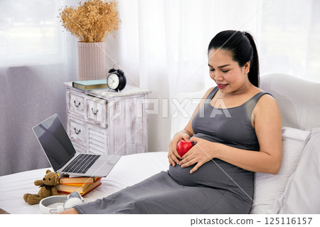 Pregnant woman in grey dress holding red heart on belly while sitting on bed next to laptop, books, and teddy bear with love, anticipation, and modern motherhood in cozy home 125116157