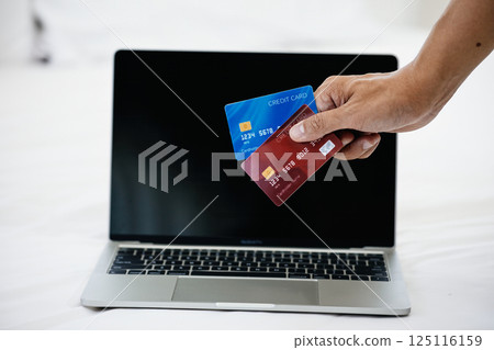 Close-up of hand holding red and blue credit cards in front of laptop screen on white bed; online payment, digital banking, and financial technology for secure e-commerce transactions 125116159