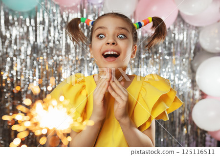 Happy funny girl in glasses and yellow holiday blouse with ponytails hairstyle on festive background with balloons looks at sparklers 125116511