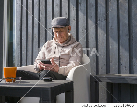 Elderly man relaxing on white chair, wearing baseball cap and glasses, listening to music through earbuds while browsing smartphone on wooden porch Elderly man relaxing on white chair, wearing baseball cap and glasses, listening to music through earbuds while browsing smartphone on wooden porch 125116600
