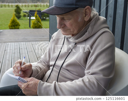 Senior writer working in sunlit terrace setting, wearing newsboy cap, jotting down thoughts while resting near steaming beverage in white armchair Senior writer working in sunlit terrace setting, wearing newsboy cap, jotting down thoughts while resting near steaming beverage in white armchair 125116601