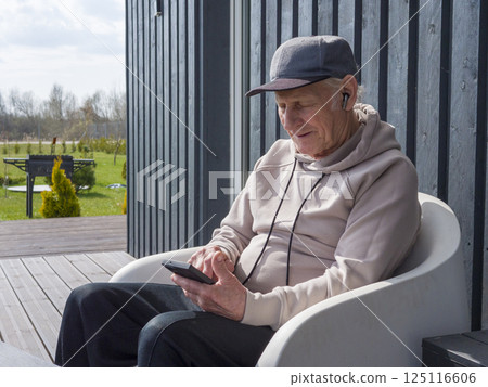 Senior male relaxing on bench, wearing baseball cap and hoodie, listening to music through wireless earphones while browsing smartphone screen Senior male relaxing on bench, wearing baseball cap and hoodie, listening to music through wireless earphones while browsing smartphone screen 125116606