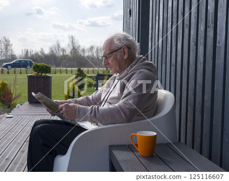 Mature man reading newspaper, wearing glasses, sitting on sleek white chair, relaxing in bright sunlight near garden, radiating calm and contentment 125116607